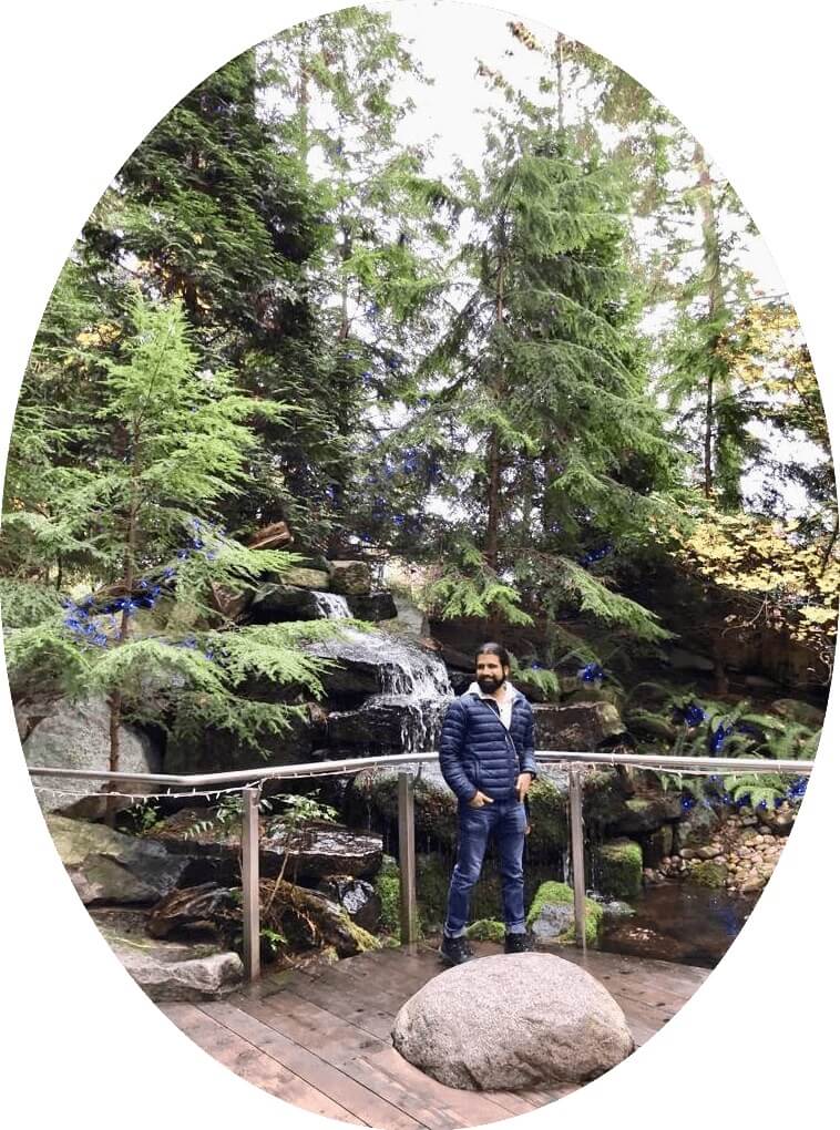In this About Me, Dr. Wadhwa is enjoying a beautiful scenery of dense tall trees in the back and a round rock in front.